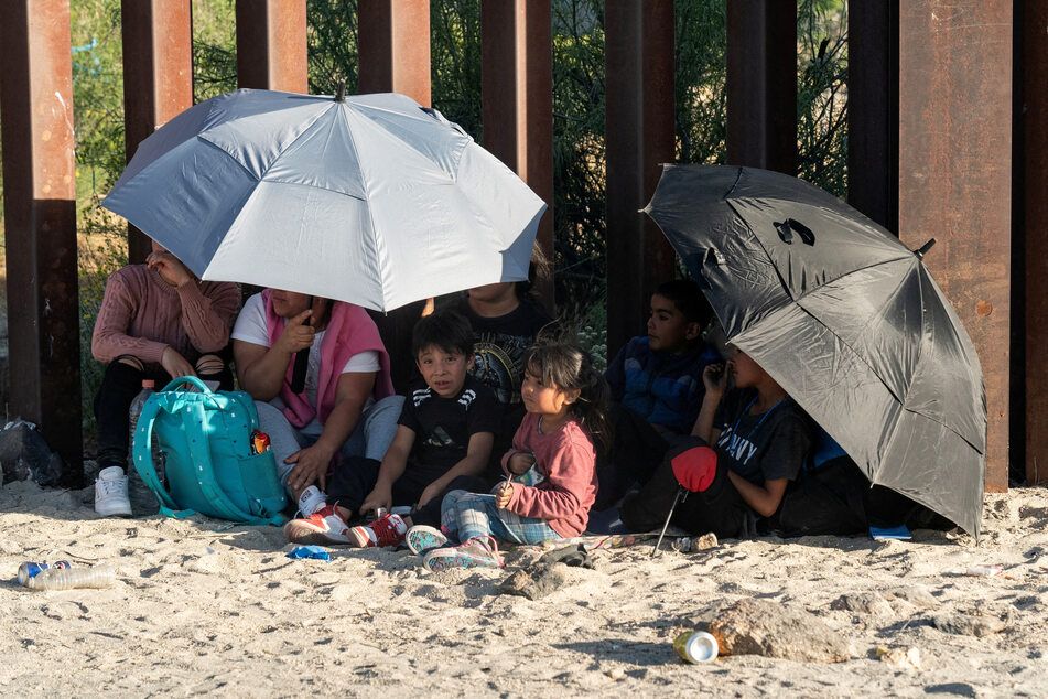 People seeking asylum shelter under umbrellas for shade while waiting at the US-Mexico border wall.