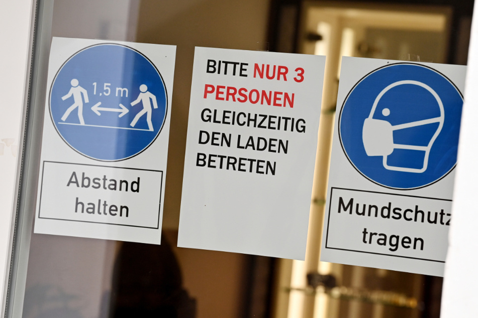 Schilder mit der Aufschrift "Abstand halten", "Bitte nur 3 Personen gleichzeitig den Laden betreten" und "Mundschutz tragen" hängen an der Tür eines Geschäftes. (Symbolbild)