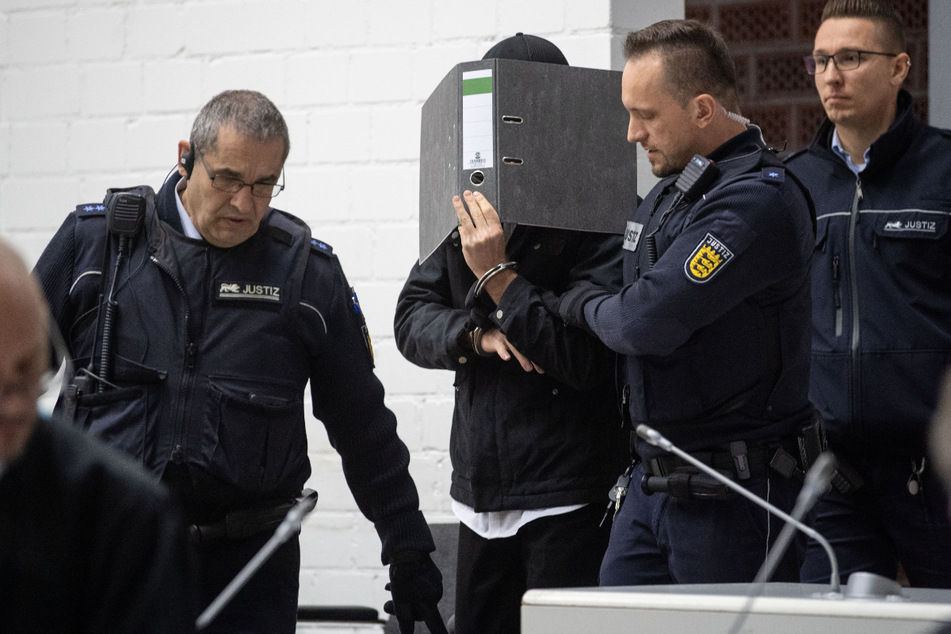Ein mutmaßlicher Islamist (m) hält sich im Gerichtssaal des Oberlandesgerichtes einen Ordner vor das Gesicht und wird von Justizbeamten an seinen Platz geführt.