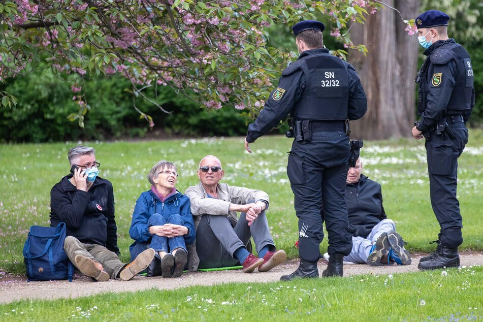 Zwei Beamte kontrollieren vier Personen, die im Großen Garten auf dem Boden sitzen.