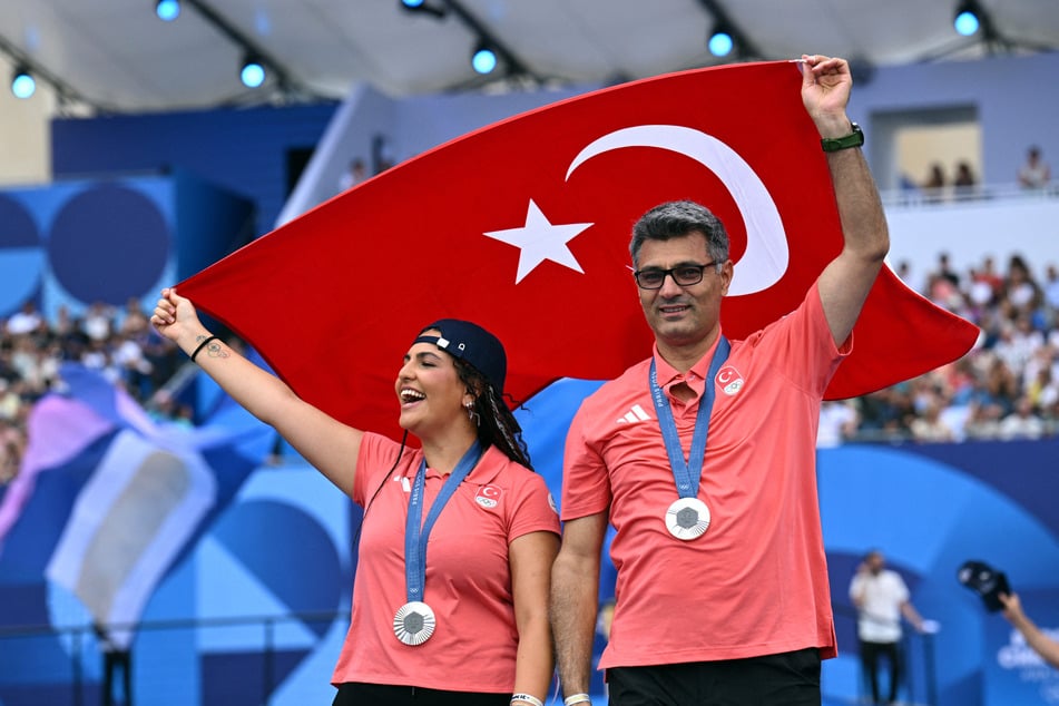 Shooting mixed silver medalists Sevval Ilayda Tarhan (l.) and Yusuf Dikec pose with the flag of Turkey during the Champions Park celebrations.