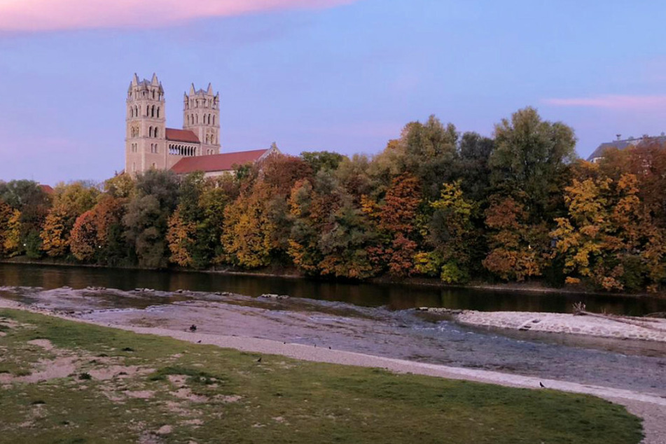 Ein farbiger Himmel zeigt sich beim Sonnenaufgang über der Isar. Im Hintergrund ist die katholische Pfarrkirche St. Maximilian zu sehen.