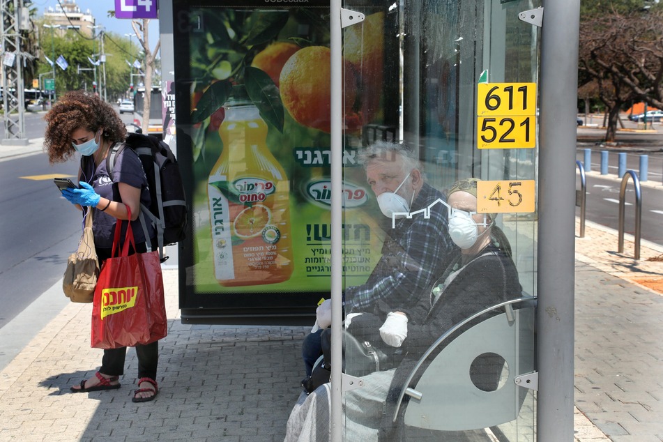 Menschen mit Mundschutz warten an einer Bushaltestelle in Tel Aviv auf den Bus. Angesichts der Ausbreitung des Coronavirus' ist auch in Israel eine Maskenpflicht verhängt worden.