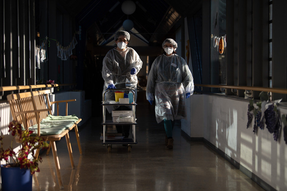 Zwei Altenpflegerinnen gehen in einem Seniorenheim mit Schutzausrüstung, die sie für die Durchführung von Antigen-Schnelltest angelegt haben, durch einen Gang.