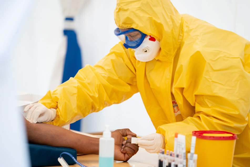 Eine Rettungssanitäterin entnimmt am frühen Morgen in Schutzkleidung in einem Zelt einem Patientin Blut.