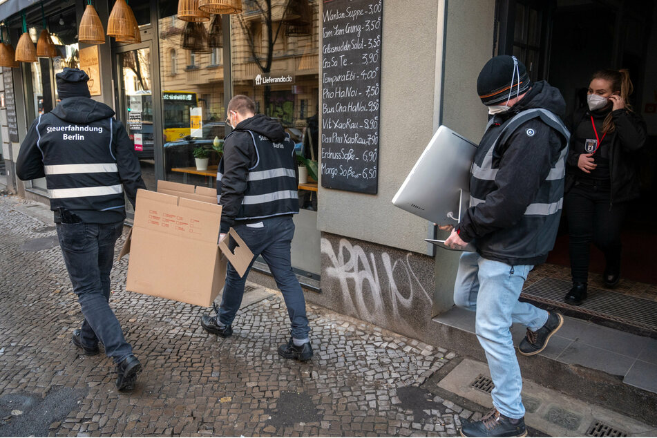 Polizisten tragen bei einer Razzia in Berlin-Neukölln eine Kiste und einen Computer aus einem Haus.