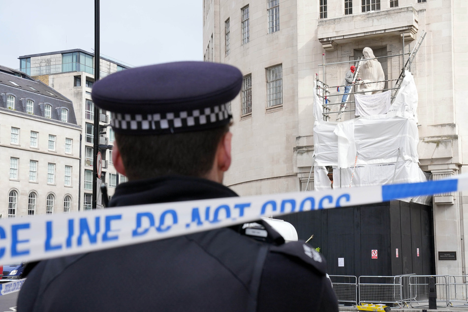 Ein Polizist sieht nur dabei zu, wie ein Mann, der eine Spiderman-Maske trägt, mit Hammer und Meißel eine Statue am BBC-Hauptquartier beschädigt.