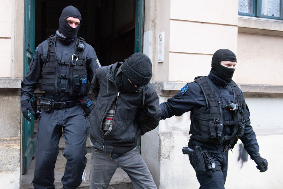 Polizisten führen bei einer Razzia einen Tatverdächtigen aus einem Haus. Nach Drohungen gegen Sachsens Ministerpräsidenten Kretschmer (46, CDU) auf Telegram durchsuchte die Polizei Mitte Dezember in Dresden mehrere Objekte.