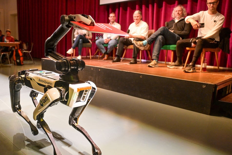 Roboterhund Spot spielt eine Rolle im Sinfoniker-Projekt, hier bringt er einen wichtigen Förderbescheid. Auf dem Podium (v.l.): Marcel Krause (Ostsächsische Sparkasse), Markus Rindt, Frank Fitzek und Moritz Lobeck, Programmleiter Musik in Hellerau.