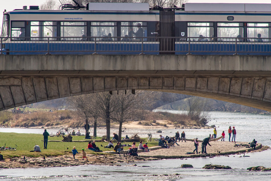 Passanten halten sich am Ufer der Isar auf, während eine Trambahn über die Reichenbachbrücke in München fährt.