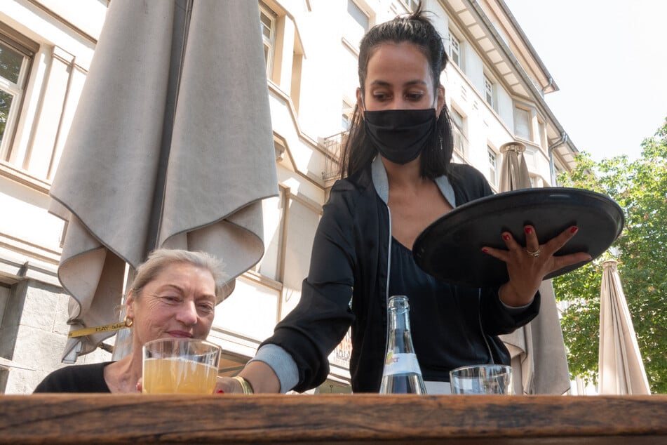 Eine Kellnerin mit Mundschutz stellt auf der Terrasse vor dem Café Kaiserbau in Stuttgart einer Kundin ein Getränk auf den Tisch.