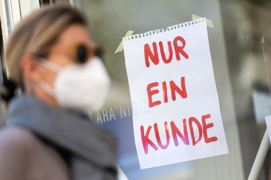 Eine Frau mit Nase-Mund-Schutzmaske geht an einem Geschäft vorbei, an dem ein Schild mit der Aufschrift "Nur ein Kunde" angebracht ist.