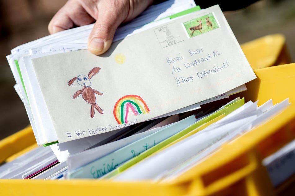 Ein Mitarbeiter des Osterpostamts hält vor dem Gemeindehaus einen Brief in den Händen, der einen gemalten Regenbogen und die Aufschrift "Wir bleiben Zuhause!" zeigt.