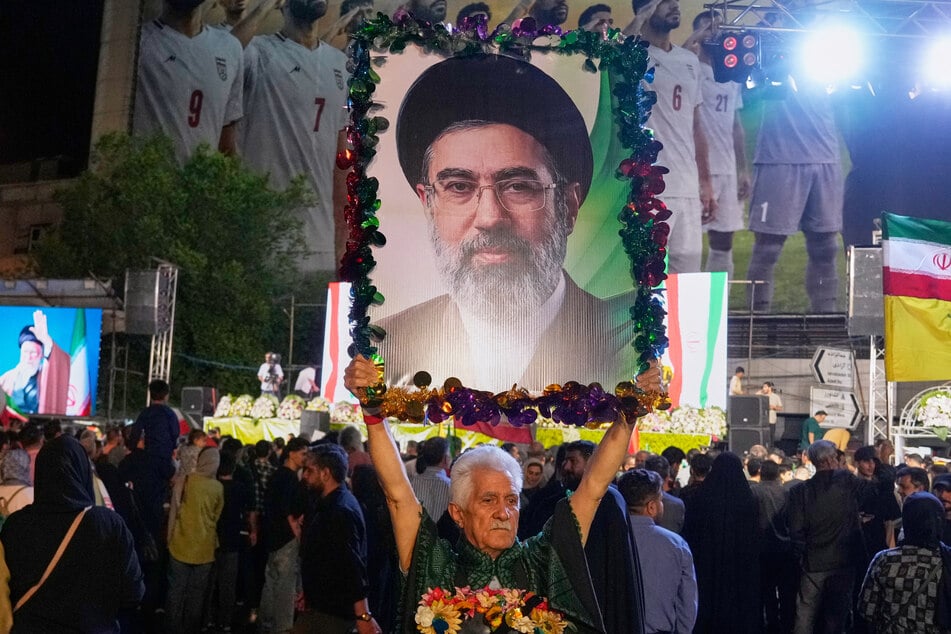 Vor dem Hintergrund eines Plakats der iranischen Fußballnationalmannschaft hält ein Mann ein Porträt des Obersten Führers Ayatollah Modschtaba Chamenei (56).