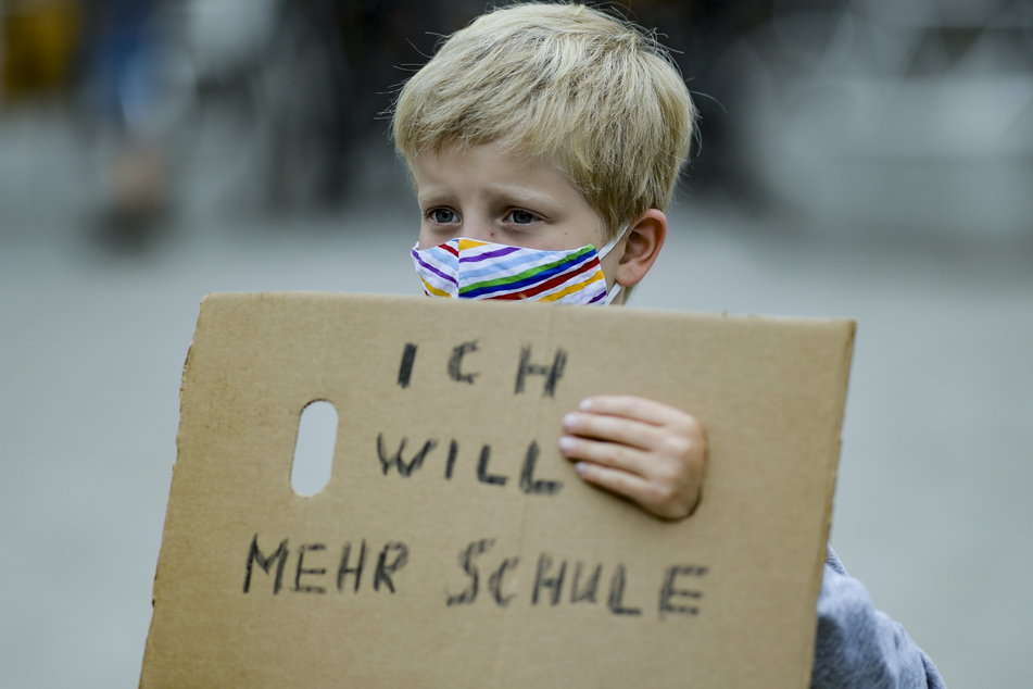 Ein Junge trägt einen bunt gestreiften Mund-Nasenschutz, während er ein Stück Pappkarton mit der Aufschrift "Ich will mehr Schule" vor sich hält.