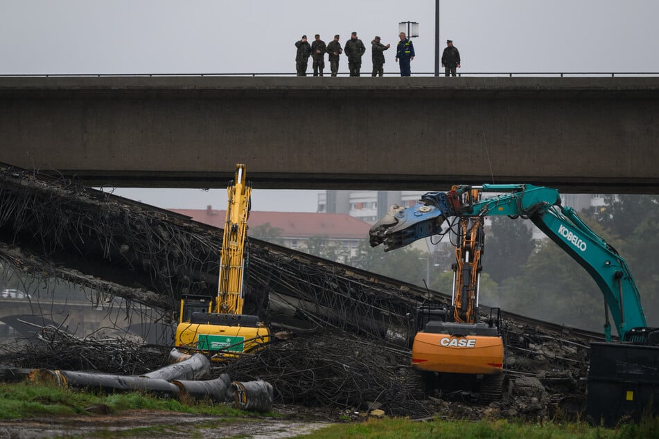 Soldaten der Bundeswehr und ein Mitarbeiter des THW beobachten wie Bagger weitere Teile der eingestürzten Carolabrücke abreißen und stehen dabei auf dem verbliebenen Teil der Brücke.