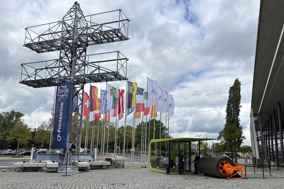 Das Mast-Exponat vor dem Dresdner Kongresszentrum. Die orange-schwarze Rolle daneben zeigt eine Behelfsstraße, die beim Leitungsbau temporär zum Einsatz kommt.