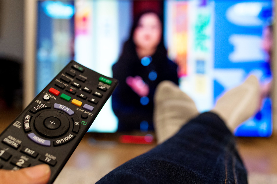 Eine Frau sitzt vor einem Fernseher und hält eine Fernbedienung in der Hand. (Symbolbild)