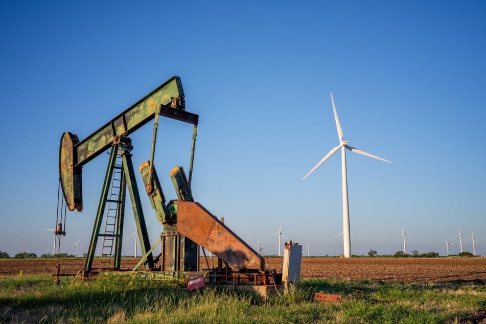 An oil pump jack operates in a field in Nolan, Texas, with a wind turbine in the background.