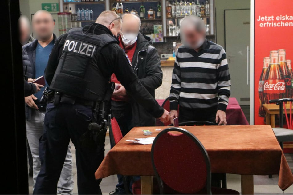 Ein Polizist diskutiert mit dem Café-Betreiber, der seine Maske noch immer nicht ordnungsgemäß trägt. Ein anderer Gast hat seinen Mund-Nasen-Schutz mittlerweile angelegt, ein Dritter verzichtet jedoch weiterhin vollständig darauf.