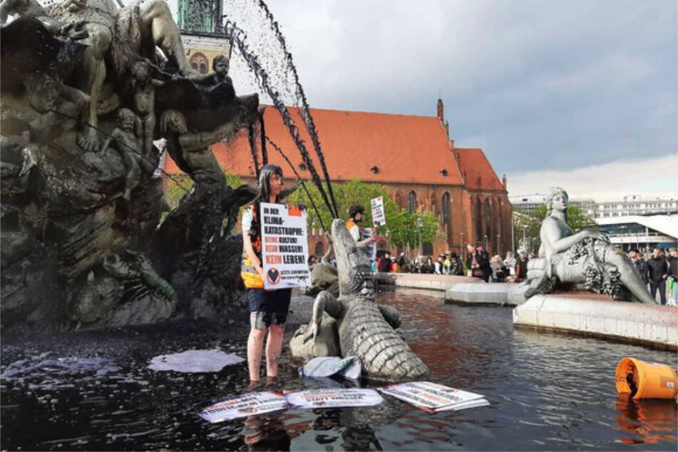 Aktivistin Judith Beadle steht im schwarz gefärbten Wasser der Brunnenanlage und hält ein Protest-Plakat hoch.