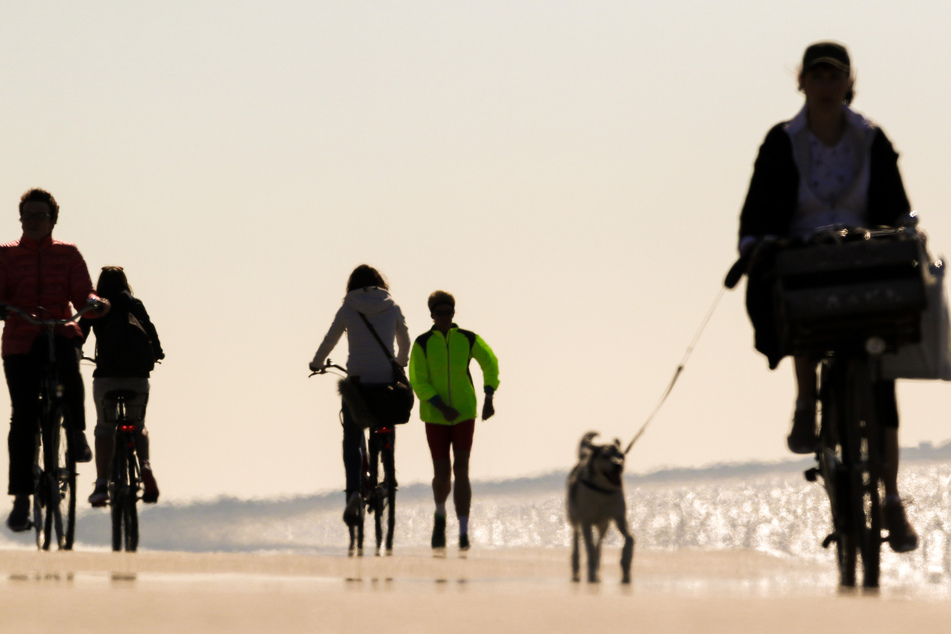 Ein Frau in einer grünen Jacke betreibt bei schönem Wetter an der Strandpromenade die Sportart Walking, während Radfahrer links und rechts an ihr vorbeifahren.