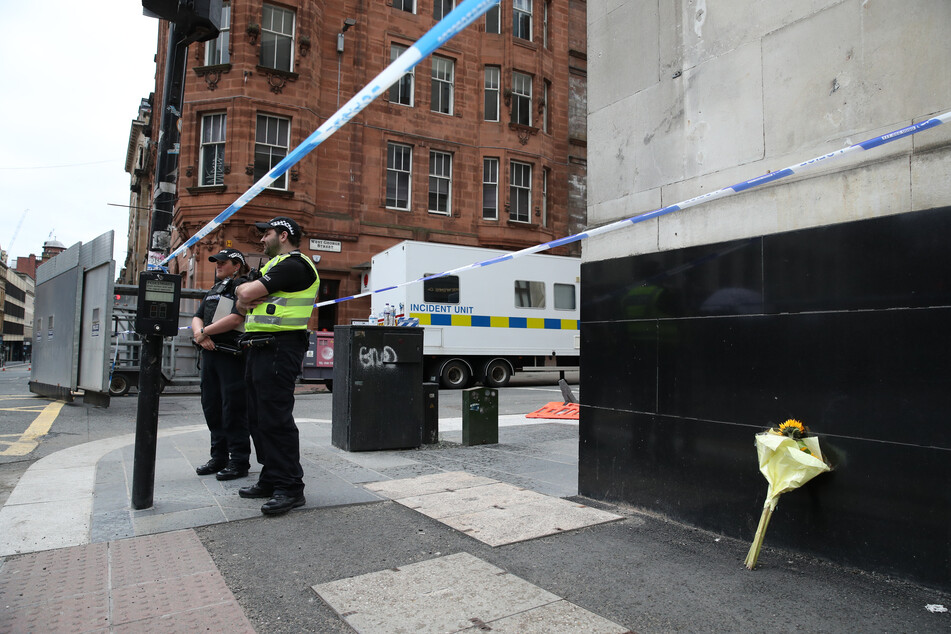 Großbritannien, Glasgow: Polizeibeamte stehen an dem abgesperrten Tatort in der West George Street, an dem ein Blumenstrauß an eine Hauswand gestellt wurde.