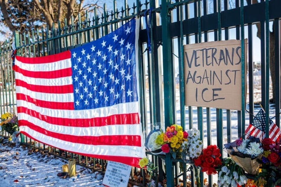 Eine US-Flagge und ein Schild mit der Aufschrift "Veterans Against ICE" hängen neben Blumen an einer Gedenkstätte zu Ehren von Alex Pretti (†37).