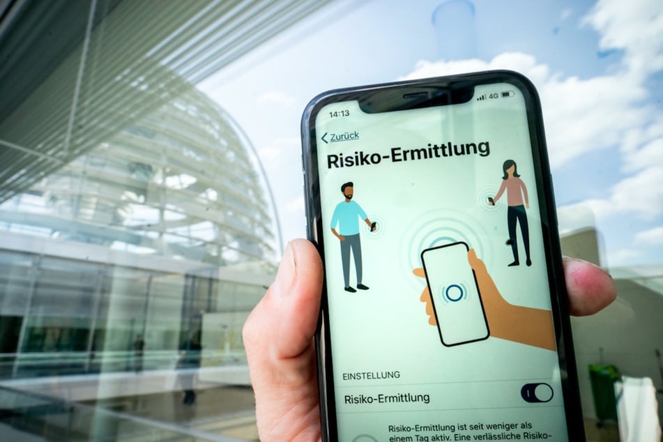 Die Corona-Warn-App mit der Seite zur Risiko-Ermittlung ist im Display eines Smartphone vor der Kuppel des Reichstags zu sehen.