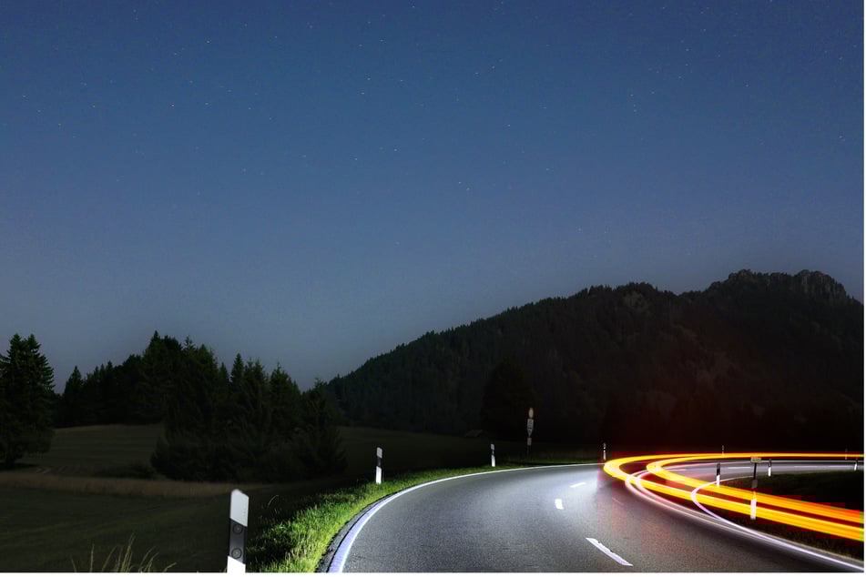Die Scheinwerfer eines Autos beleuchten eine Gebirgsstraße. Vom Sternenhimmel sieht man allerdings wegen des hellen Lichts des abnehmenden Mondes nicht so viel.