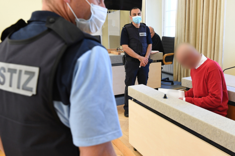 Der angeklagte Supermarkt-Erpresser (rechts) sitzt umgeben von Justizbeamten auf der Anklagebank und liest ein Dokument.