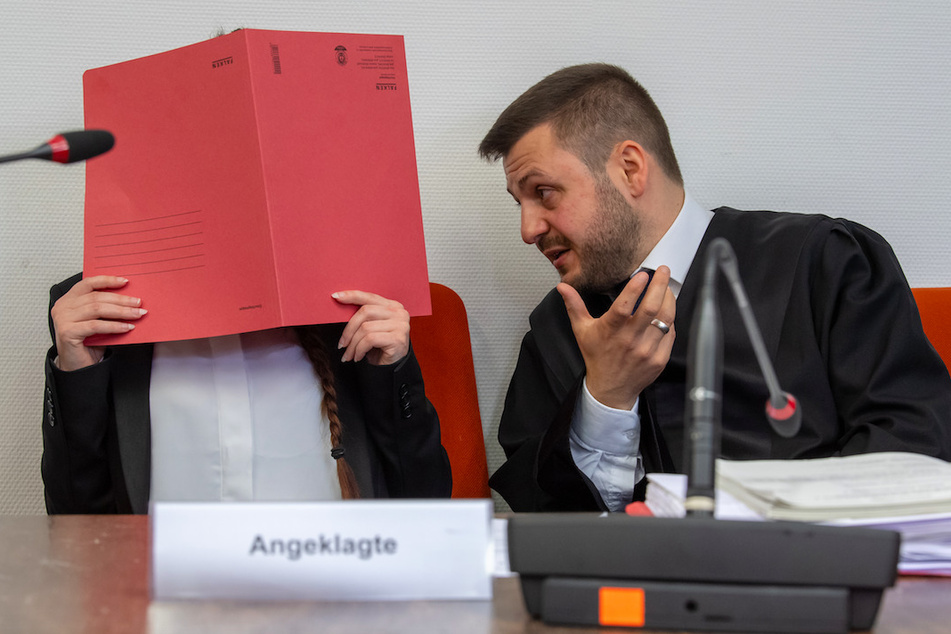 Die Angeklagte Jennifer W. hält auf ihrem Sitz im Gerichtssaals einen roten Aktendeckel vor ihr Gesicht. Rechts spricht ihr Anwalt Ali Aydin mit ihr.