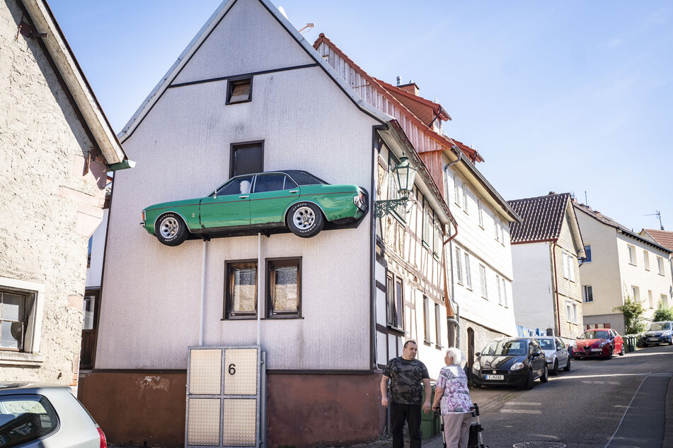 Matthias Hoffmann (u.) steht vor seinem Haus, an dessen Fassade er die linke Seite eines alten, längs aufgeschnittenen Ford Granada auf zwei Stützen angebracht hat.