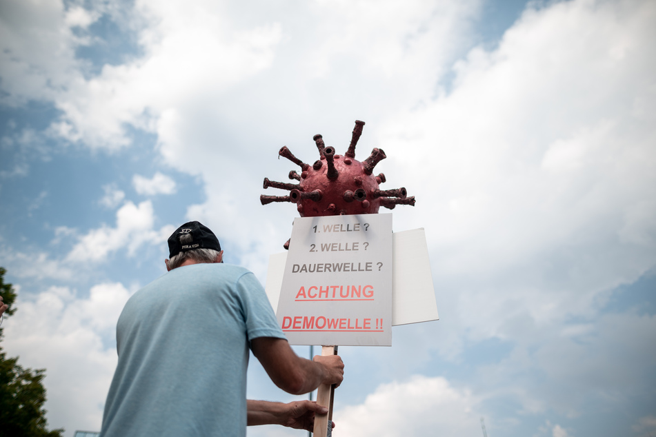 Ein Teilnehmer der Demonstration hält ein Schild mit einem Coronavirus-Symbol auf dem "1.Welle, 2. Welle, Dauerwelle, Achtung Demowelle" steht.