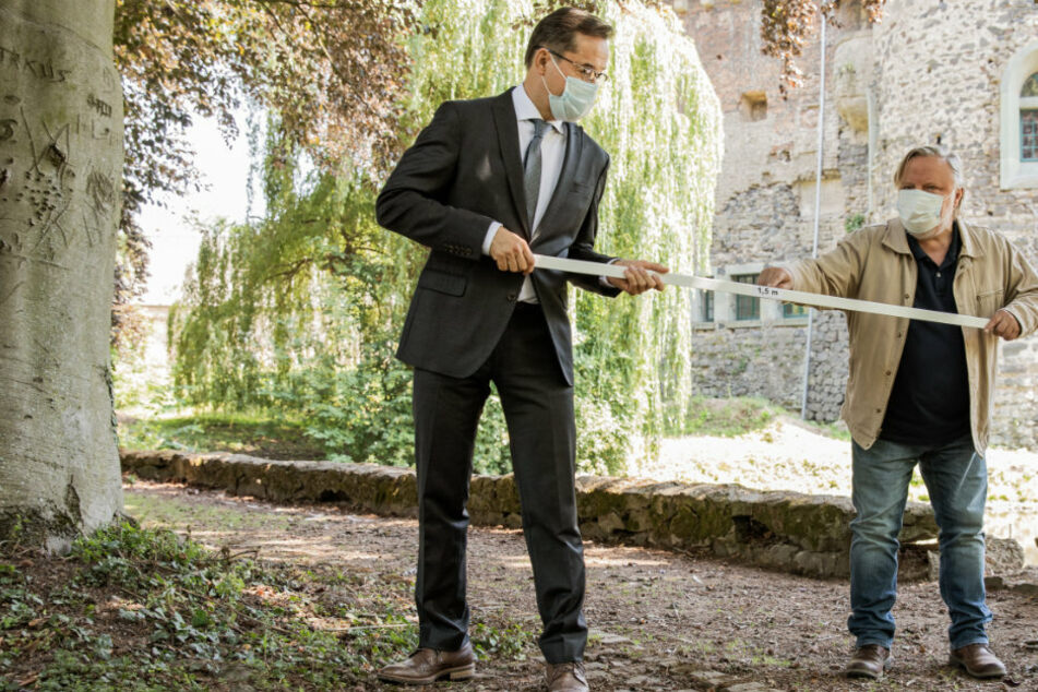 Jan Josef Liefers (l) und Axel Prahl (r) stehen bei Dreharbeiten zum neuen "Tatort" aus Münster vor Schloß Hülchrath und zeigen mit Hilfe einer 1,5 Meter langen Latte den Mindestabstand.