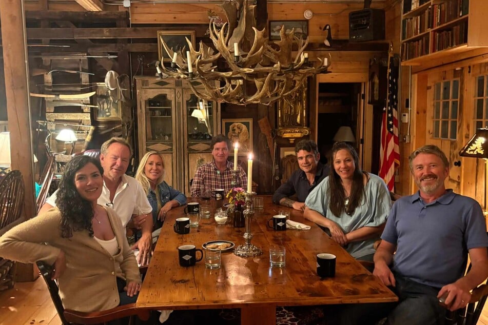 Marjorie Taylor Greene (3rd from L), Tucker Carlson (c), Joe Kent (3rd from R), and Rep. Thomas Massie (far r) sit at a hunting cabin along with their family members.