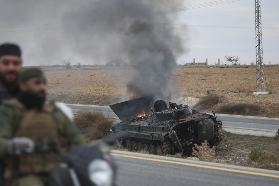 Syrian opposition fighters drive past a burning government armored vehicle south of Hama, Syria.