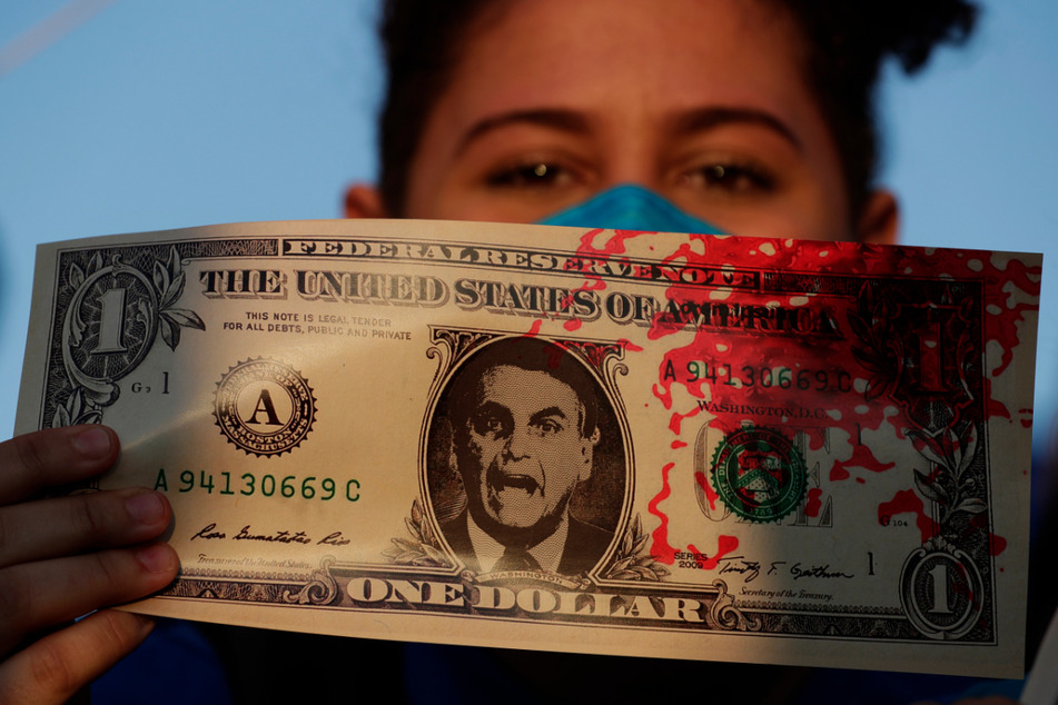Eine Frau hält eine Kopie eines US-Dollar-Scheins, auf dem ein Bild des brasilianischen Präsidenten Jair Bolsonaro sowie rote Farbe, die das Blut von Covid-19-Opfern symbolisiert, zu sehen ist.