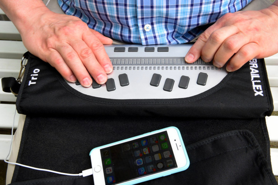 Ein Mann Arbeitet mit einer Braillezeile und einem Smartphone mit Kopfhörern. Die Braillezeile ist ein Ausgabegerät für Blinde, welches Texte in Brailleschrift umwandelt und mit dem Tastsinn erfühlt werden können.