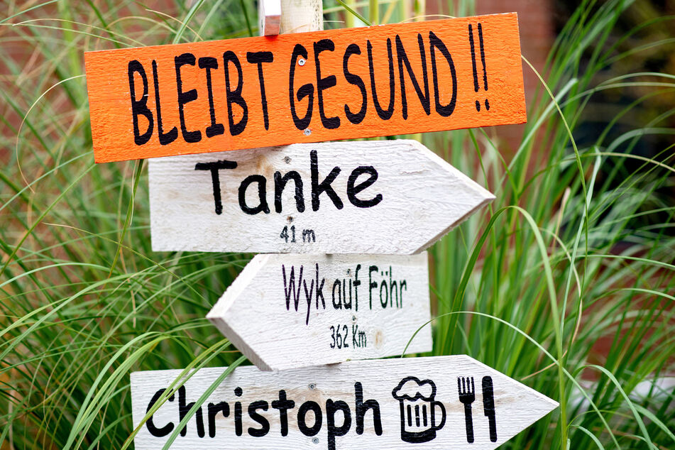 Ein Holzschild mit der Aufschrift "Bleibt gesund!" hängt an einem Wegweiser, den eine Familie vor ihrem Haus aufgestellt hat.
