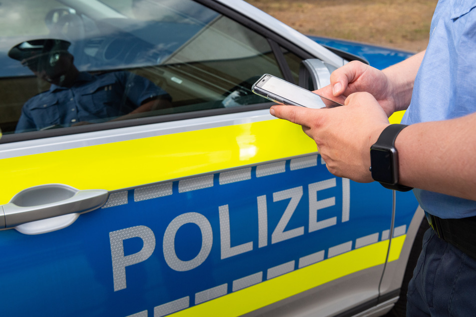 Ein Polizist steht vor seinem Wagen mit einem Smartphone in der Hand (Symbolbild).
