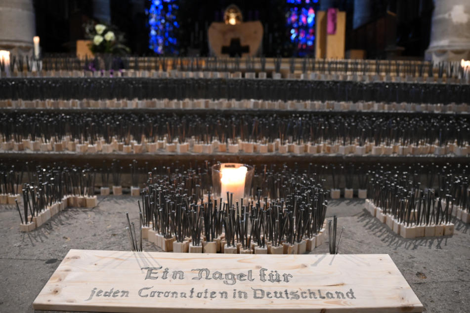 Nägel stehen im Heilig-Kreuz-Münster in einem Kunstwerk aus 14434 Nägeln. Im Vordergrund liegt ein Schild, auf dem "Ein Nagel für jeden Coronatoten in Deutschland" steht.