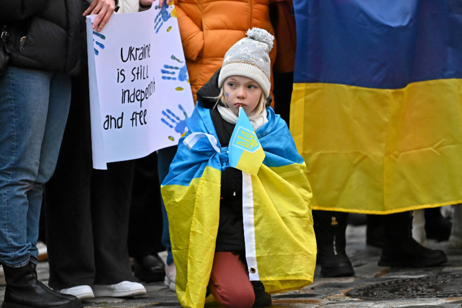 Ein kleines, in eine ukrainische Flagge gehülltes Mädchen, kniet auf den kalten Steinen des Neumarkts.