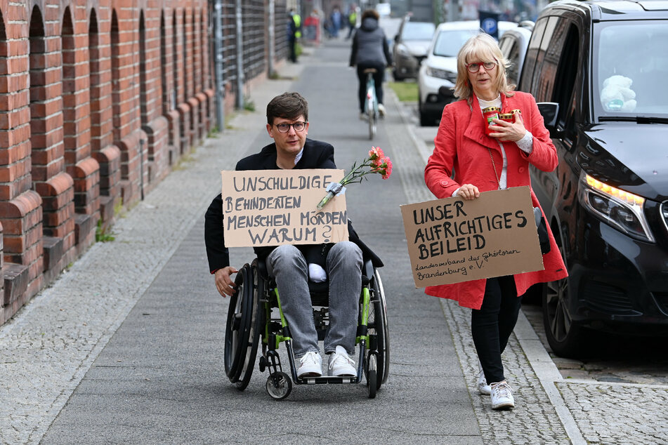 Eine Frau und ein Mann im Rollstuhl gehen mit Plakaten "Unschuldige behinderte Menschen töten. Warum?" und "Unser aufrichtiges Beileid" zum Eingang der Einrichtung des diakonischen Anbieters Oberlinhaus.