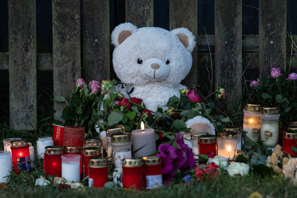 Kerzen, Blumen und ein großer Plüschteddy sind vor einem Zaun am Fundort zu sehen, wo die Leiche einer vermissten 14-Jährigen aus Nordhessen in einem Waldstück gefunden wurde.