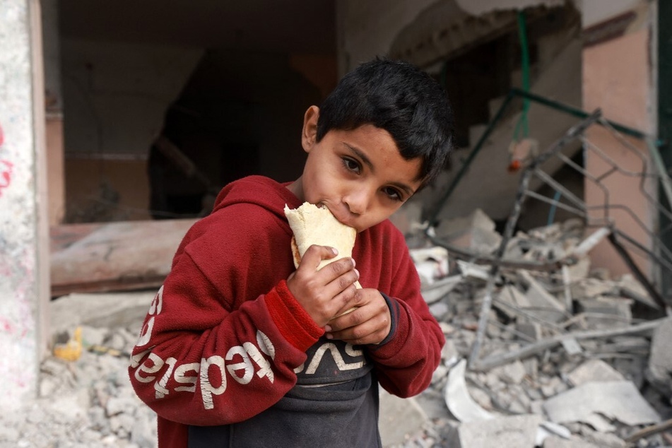 A Palestinian child eats a sandwich as he stands in front of a building damaged by Israeli bombardment in Rafah in the southern Gaza Strip on February 25, 2024.