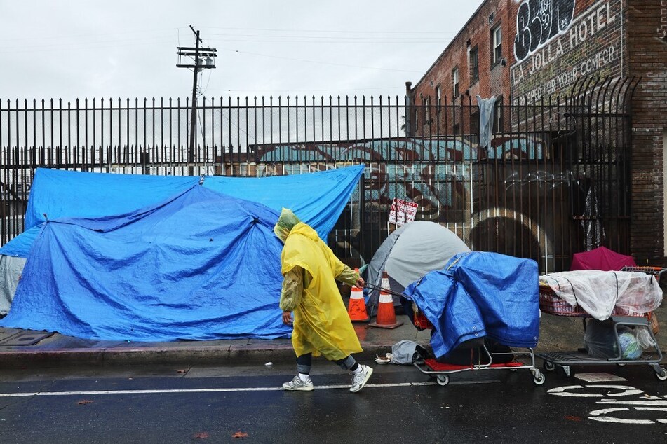 A person walks with carts in the rain near an encampment of unhoused people in Skid Row, Los Angeles, California.