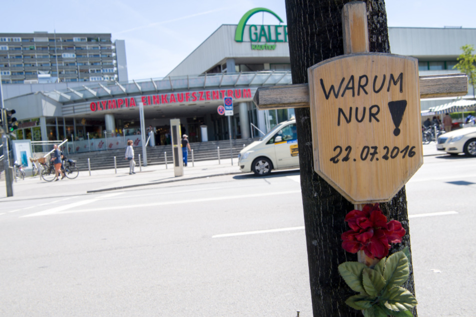 Ein Holzkreuz mit einem Schild und der Aufschrift "Warum nur! - 22.07.2016" hängt vor dem Eingang zum Olympia-Einkaufszentrum (OEZ). (Archiv)
