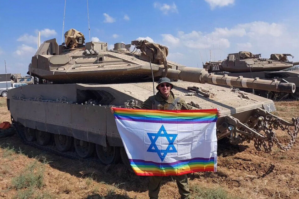 Yoav Atzmoni poses in front of an tank holding a rainbow-edged Israeli flag.
