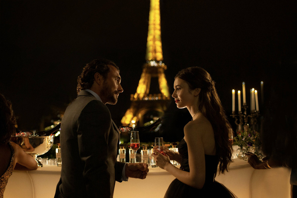 William Abadie (r) and Lily Collings (l) share a drink while overlooking the Eiffel tower, playing Antoine Lambert and Emily Cooper in season one of Emily in Paris.